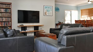 A living room with a flat-screen TV, leather sofas, a bookshelf, and a dining area with wooden chairs and a table.