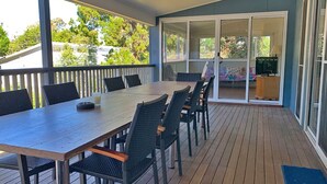 A covered outdoor area with a wooden deck, a long table with chairs, and a sliding glass door leading to a balcony.