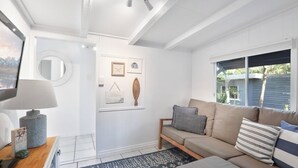 A well-lit living room with a beige sofa, a coffee table, and a TV. There are framed pictures on the wall and a window with blinds.