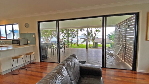 A living room with a leather sofa, a kitchen area with a microwave, and a sliding glass door leading to an outdoor deck with a view of the ocean.