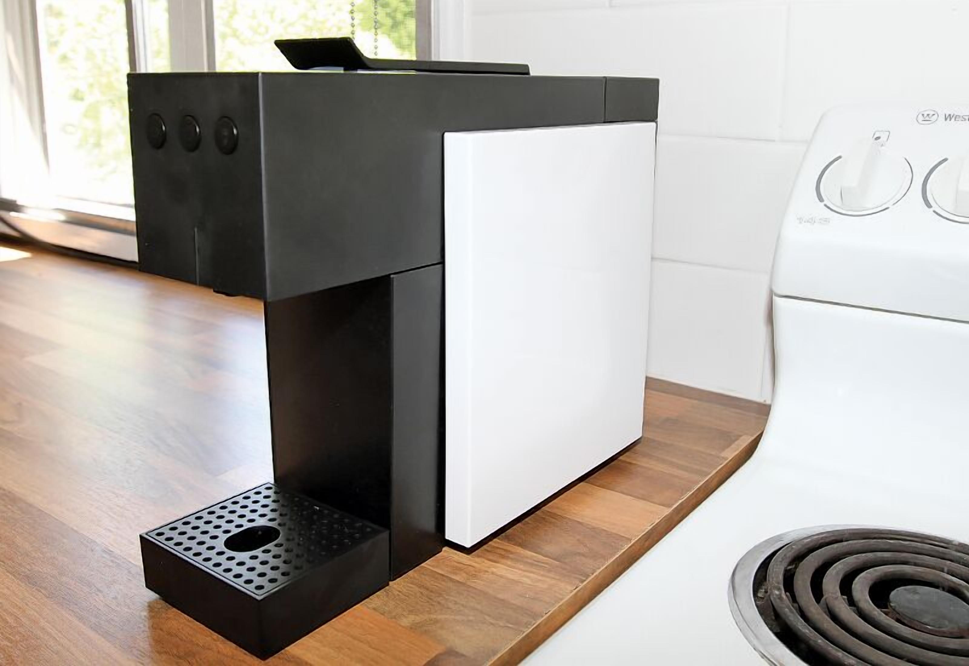 Una cafetera compacta y moderna con diseño en blanco y negro, sobre una encimera de madera, junto a un electrodoméstico blanco, probablemente una lavadora.