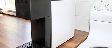 Una cafetera compacta y moderna con diseño en blanco y negro, sobre una encimera de madera, junto a un electrodoméstico blanco, probablemente una lavadora.