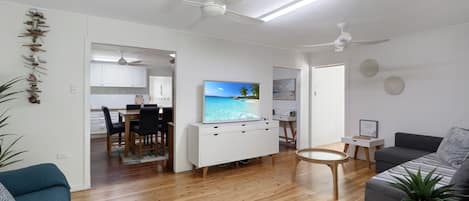 Una sala de estar moderna con piso de madera, un sofá gris, un área de comedor con televisor y ventiladores de techo.
