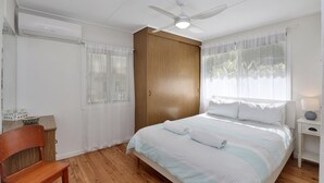 A bedroom with a bed, a wooden wardrobe, a ceiling fan, and a window with sheer curtains. - Beach Shack From Yesteryear With All The Modern Comforts (Amity Point)
