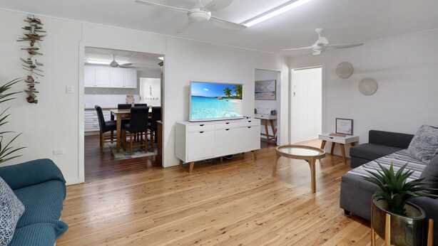 A modern living room with wooden flooring, a grey sofa, a dining area with a TV, and ceiling fans. - Beach Shack From Yesteryear With All The Modern Comforts (Amity Point)