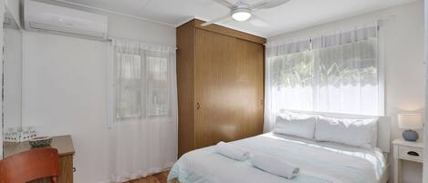 A bedroom with a bed, a wooden wardrobe, a ceiling fan, and a window with sheer curtains.