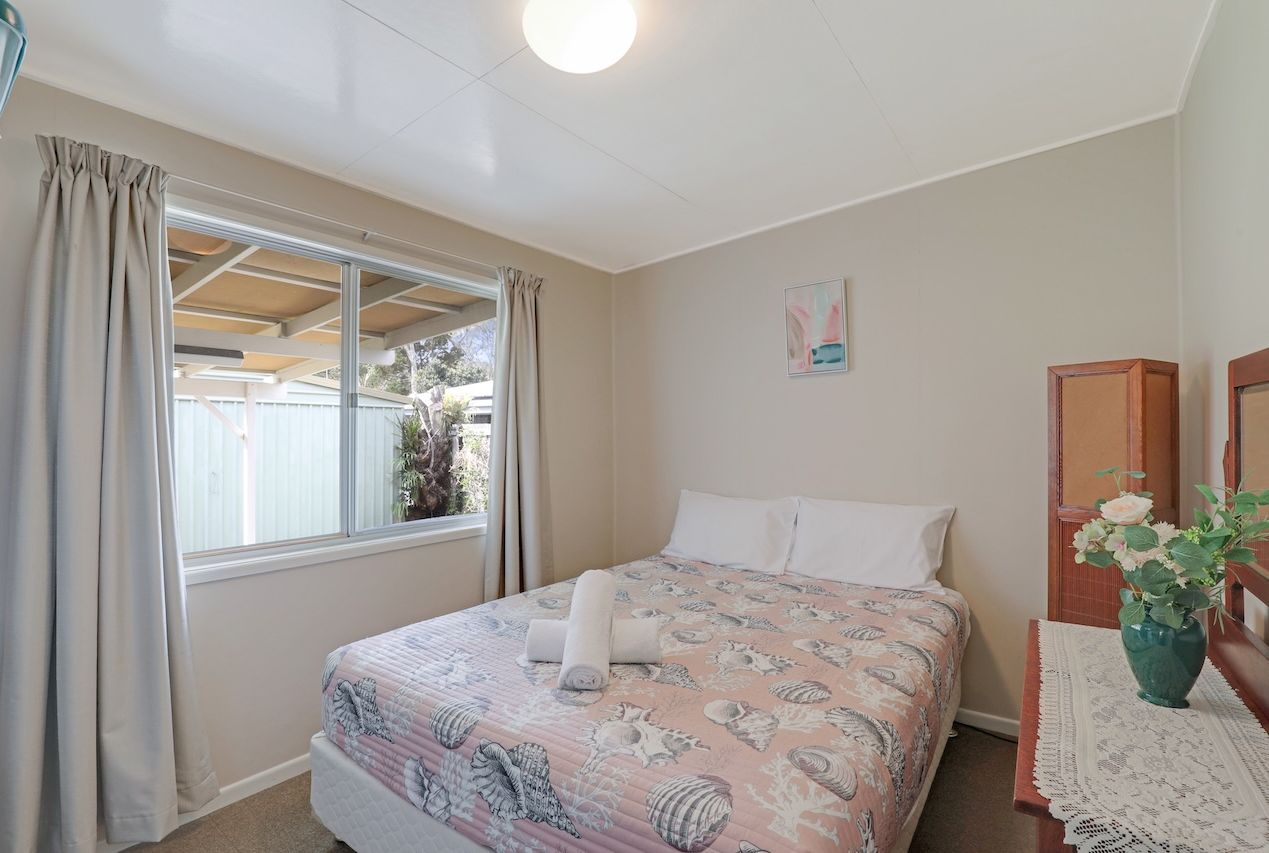 A bedroom with a bed, curtains, a window, a wooden headboard, a bedside table with a vase, and a wall-mounted picture.