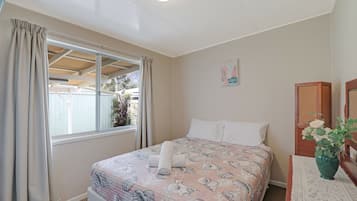 Un dormitorio con cama, cortinas, ventana, cabecera de madera, mesita de noche con un florero y un cuadro colgado en la pared.