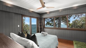 Een slaapkamer met een bed, een houten bureau, een plafondventilator en grote ramen met uitzicht op zee.