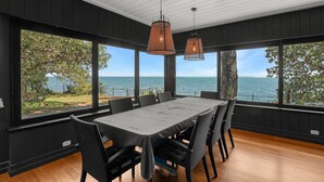 Een eethoek met een tafel en stoelen, grote ramen met uitzicht op zee, en hangende pendantlampen.