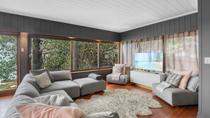 Een moderne woonkamer met een grijze bank, een zacht wit vloerkleed en grote ramen met uitzicht op bomen.