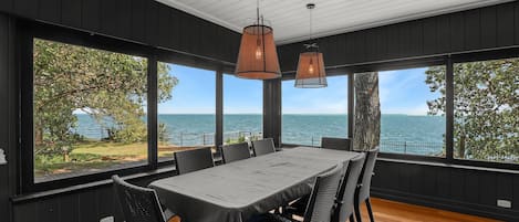 A dining area with a table and chairs, large windows with a view of the sea, and hanging pendant lights.