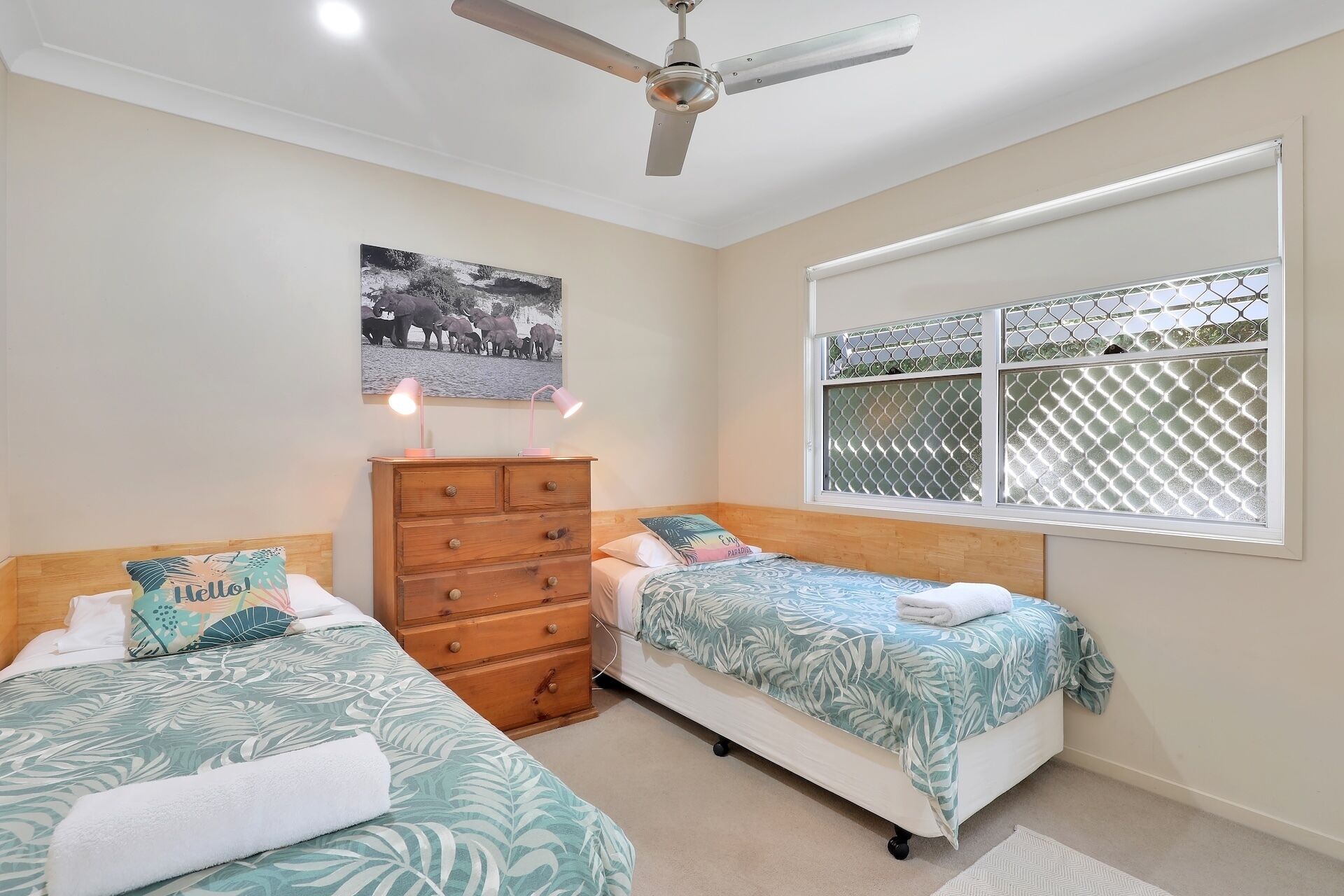 A bedroom with two beds, a wooden dresser, a ceiling fan, a window with a lattice design, and a wall-mounted picture.
