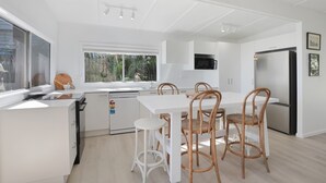 Une cuisine moderne avec des meubles blancs, un lave-vaisselle encastré, un grand réfrigérateur et un coin repas doté de tables blanches et de chaises en osier.