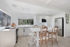 Une cuisine moderne avec des meubles blancs, un lave-vaisselle encastré, un grand réfrigérateur et un coin repas doté de tables blanches et de chaises en osier.
