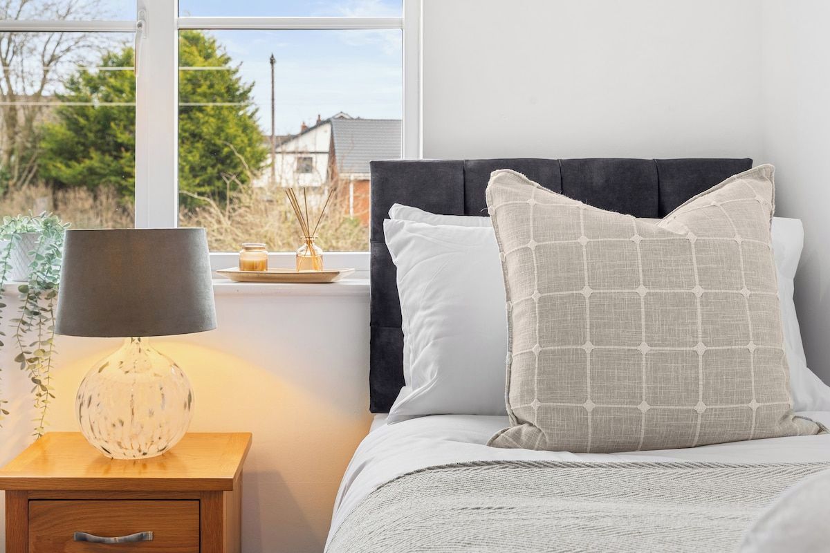 A bedroom with a bed, a bedside table with a lamp, and a window with a view of trees and houses.