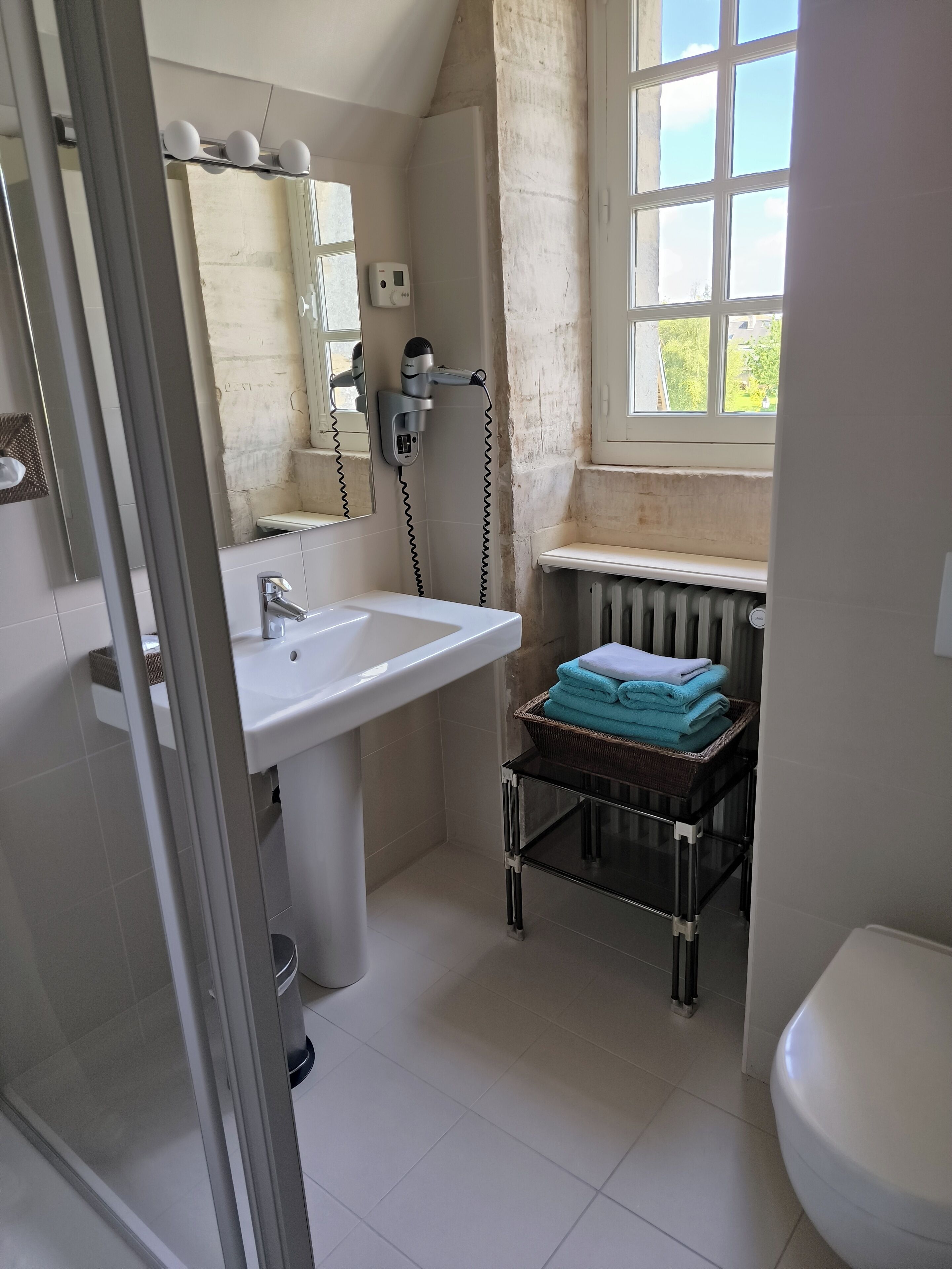 Une salle de bain avec un lavabo blanc, un miroir, un sèche-cheveux, un radiateur et une fenêtre donnant sur un paysage verdoyant.