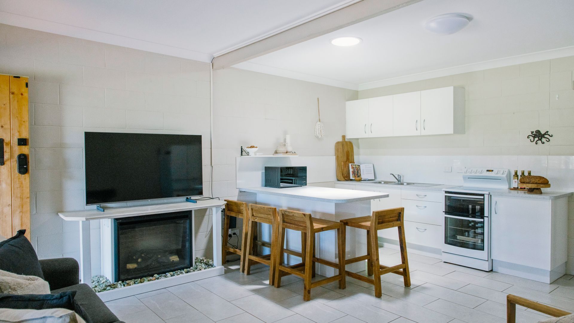 A modern kitchen with white cabinets, a built-in oven, a microwave, and a large flat-screen TV. There is a wooden dining table with chairs and a fireplace with a stone surround.