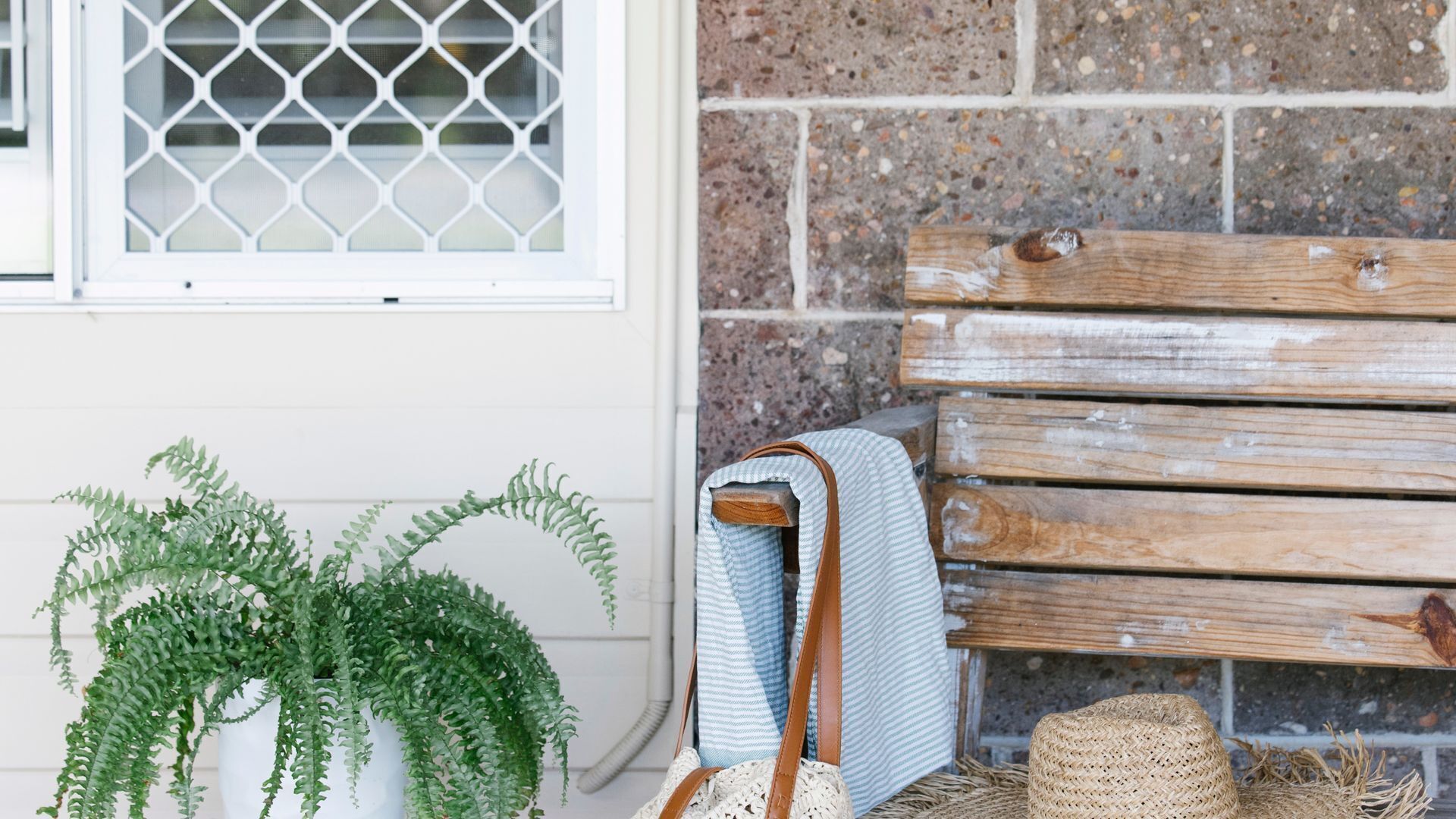 A wooden bench, a woven basket, and a potted fern plant against a brick wall.