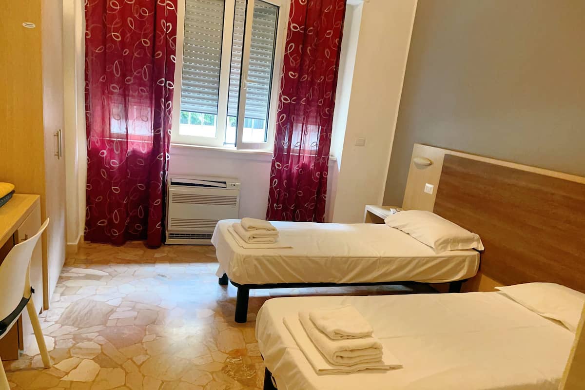 A hotel room with two single beds, a desk, a chair, a heater, and a window with curtains.