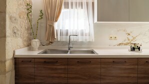 A kitchen with a marble backsplash, a double sink, wooden cabinets, and a window with sheer curtains. - Mareu Stonehouse in the Cretan countryside (Stironas)