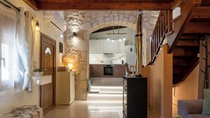 A spacious living area with a stone wall, wooden ceiling, and a view into the kitchen. - Mareu Stonehouse in the Cretan countryside (Stironas)