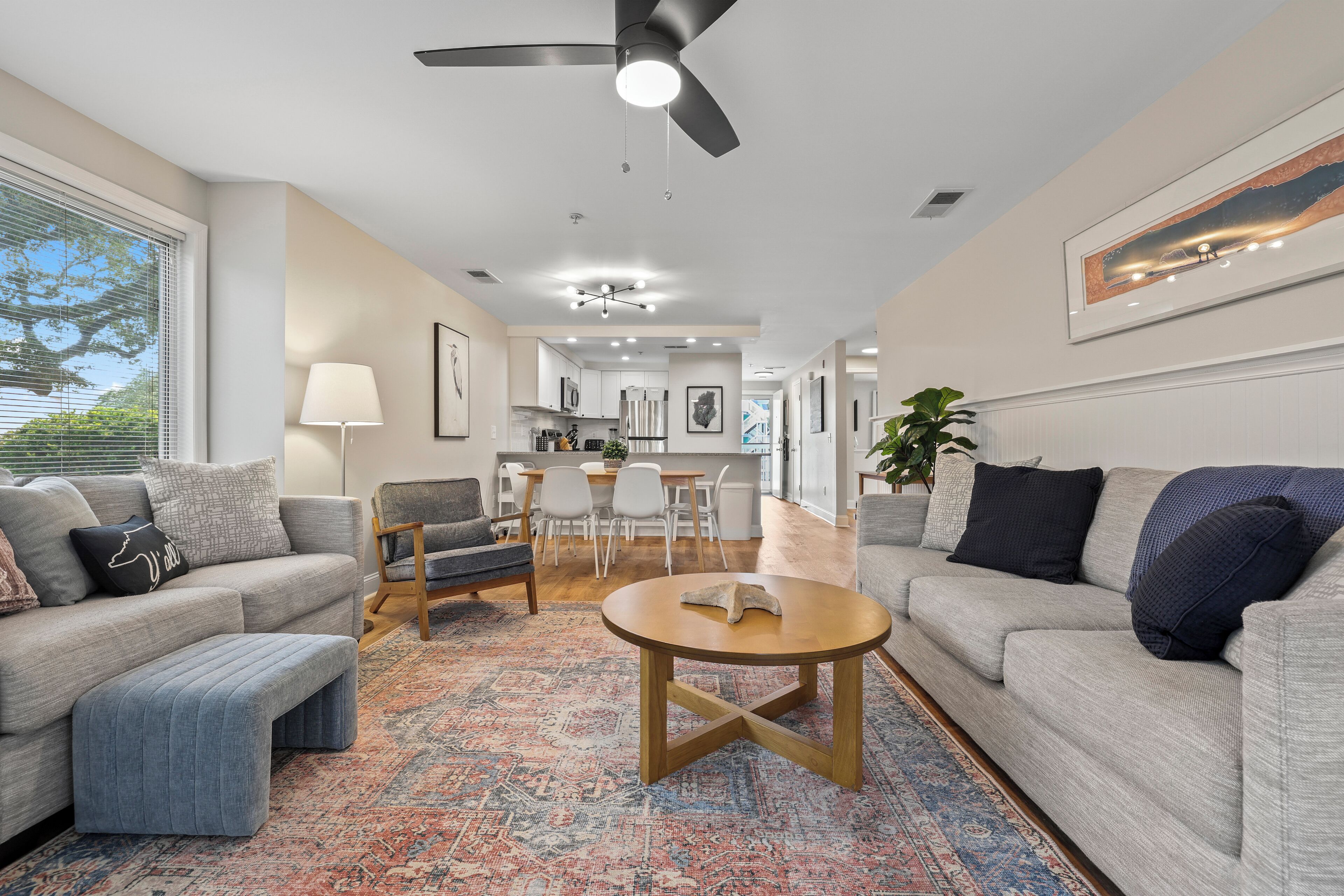 A modern living room with a grey sofa, ottoman, and coffee table, a dining area with chairs and a table, and a kitchen in the background.