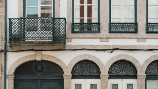 A building with a large arched entrance, a balcony with a metal railing, and a row of windows with shutters.