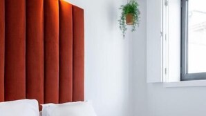 A modern hotel room with a large bed, white bedding, a red accent wall, a hanging plant, and a window with natural light.