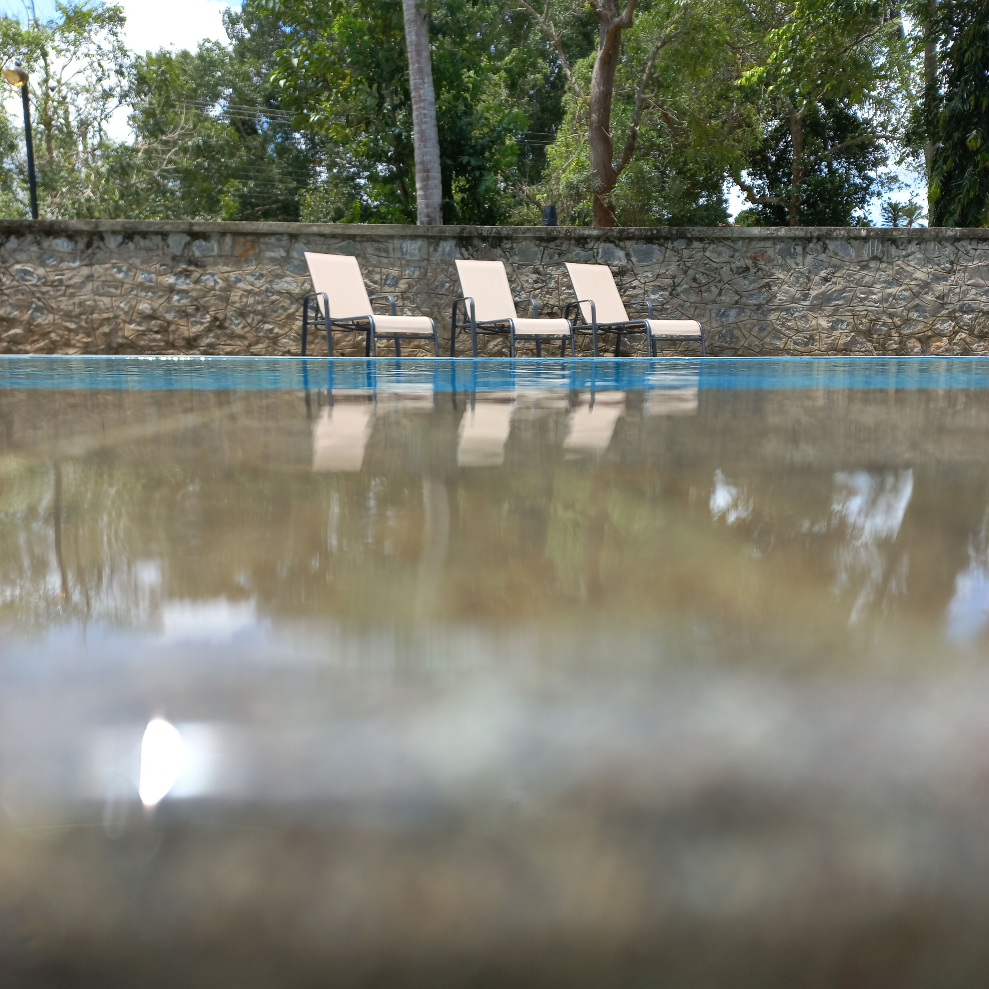 Una piscina con tres sillas de descanso y un muro de piedra al fondo.