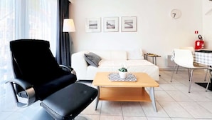 Ein modernes Wohnzimmer mit einem schwarzen Sessel, einem weißen Sofa, einem Holztisch und einem kleinen Tisch mit kariertem Tischläufer. An der Wand hängen drei gerahmte Bilder, eine Lampe und ein Feuerlöscher.