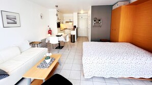Ein helles, modernes Zimmer mit einem weißen Sofa, einem Holztisch mit Tablett, einem Bett mit floralem Überwurf, einer Küchenzeile im Hintergrund und einer grauen Wand mit Regal.