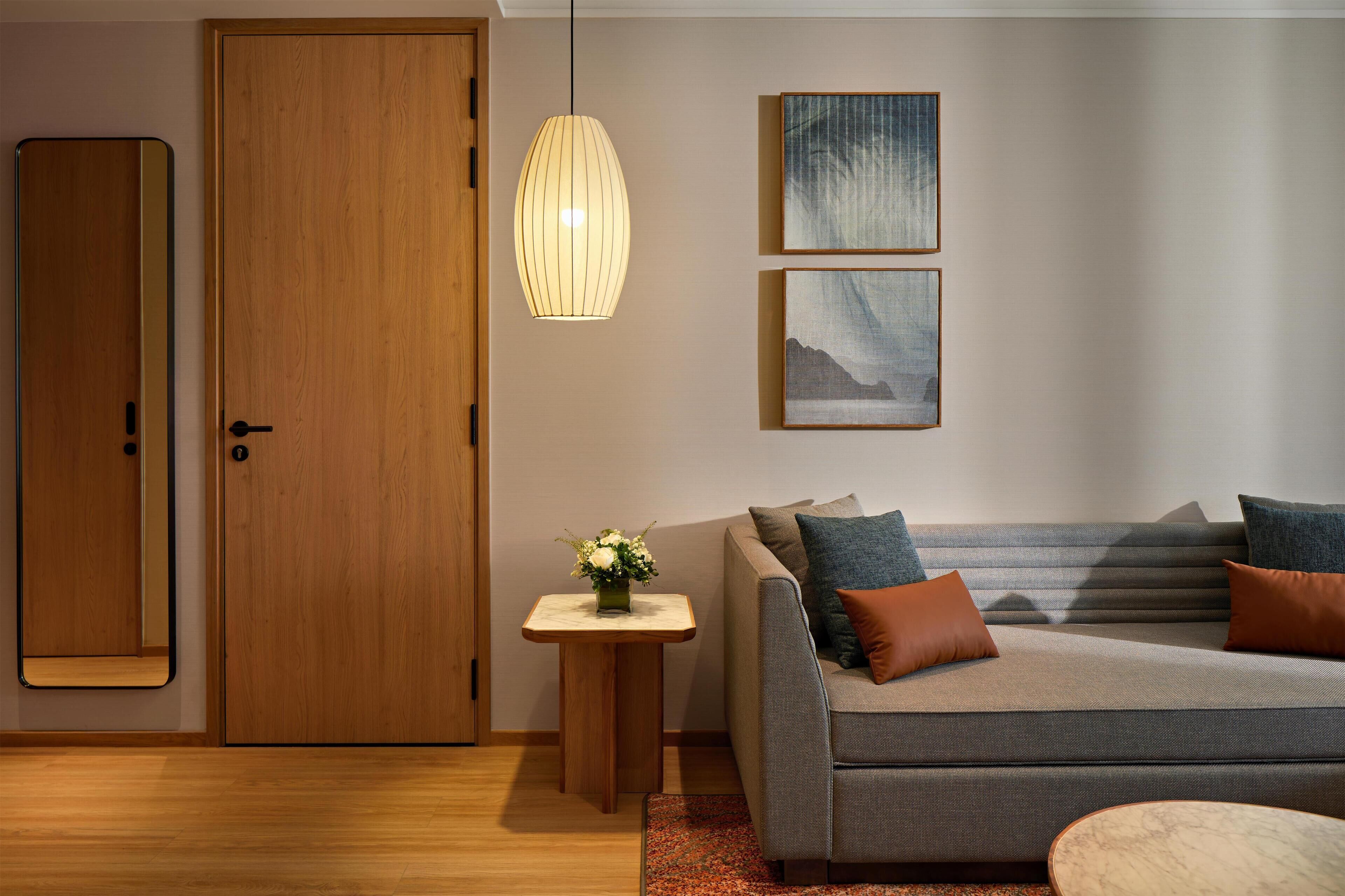 A modern living room with a wooden door, a sofa with cushions, a small table with a vase, and two framed artworks on the wall.