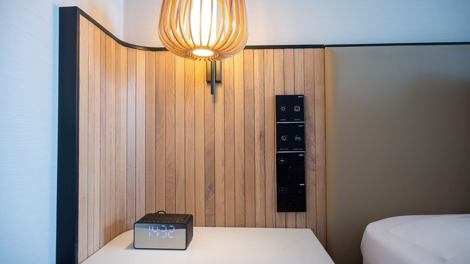 A hotel room with a wooden panel wall, a bedside table with a digital clock, and a wall-mounted control panel with multiple buttons.