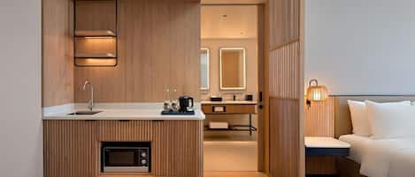 A modern hotel room with a wooden kitchenette, a bed, and a bathroom with a mirror and lights.