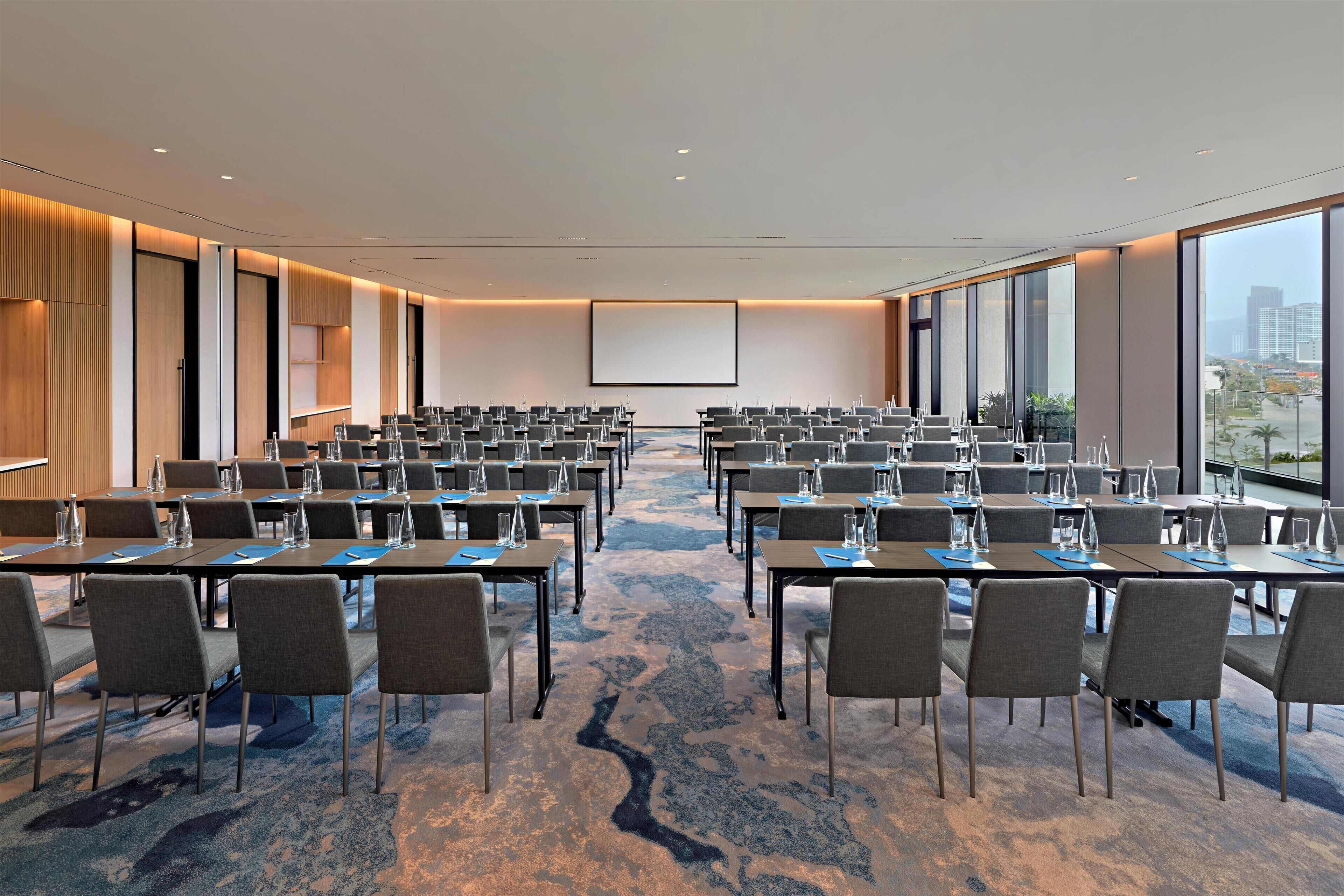 Un salón de conferencias con filas de mesas y sillas, una pantalla grande y piso alfombrado.