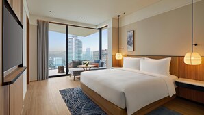 Une chambre d’hôtel moderne avec un grand lit, une télévision, un balcon offrant une vue sur la ville et une petite table agrémentée d’un vase de fleurs.
