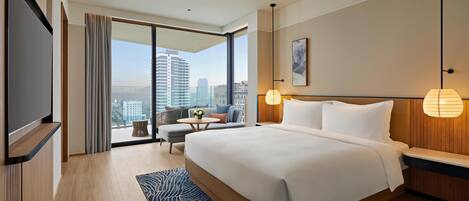A modern hotel room with a large bed, a TV, a balcony with a city view, and a small table with a flower vase.