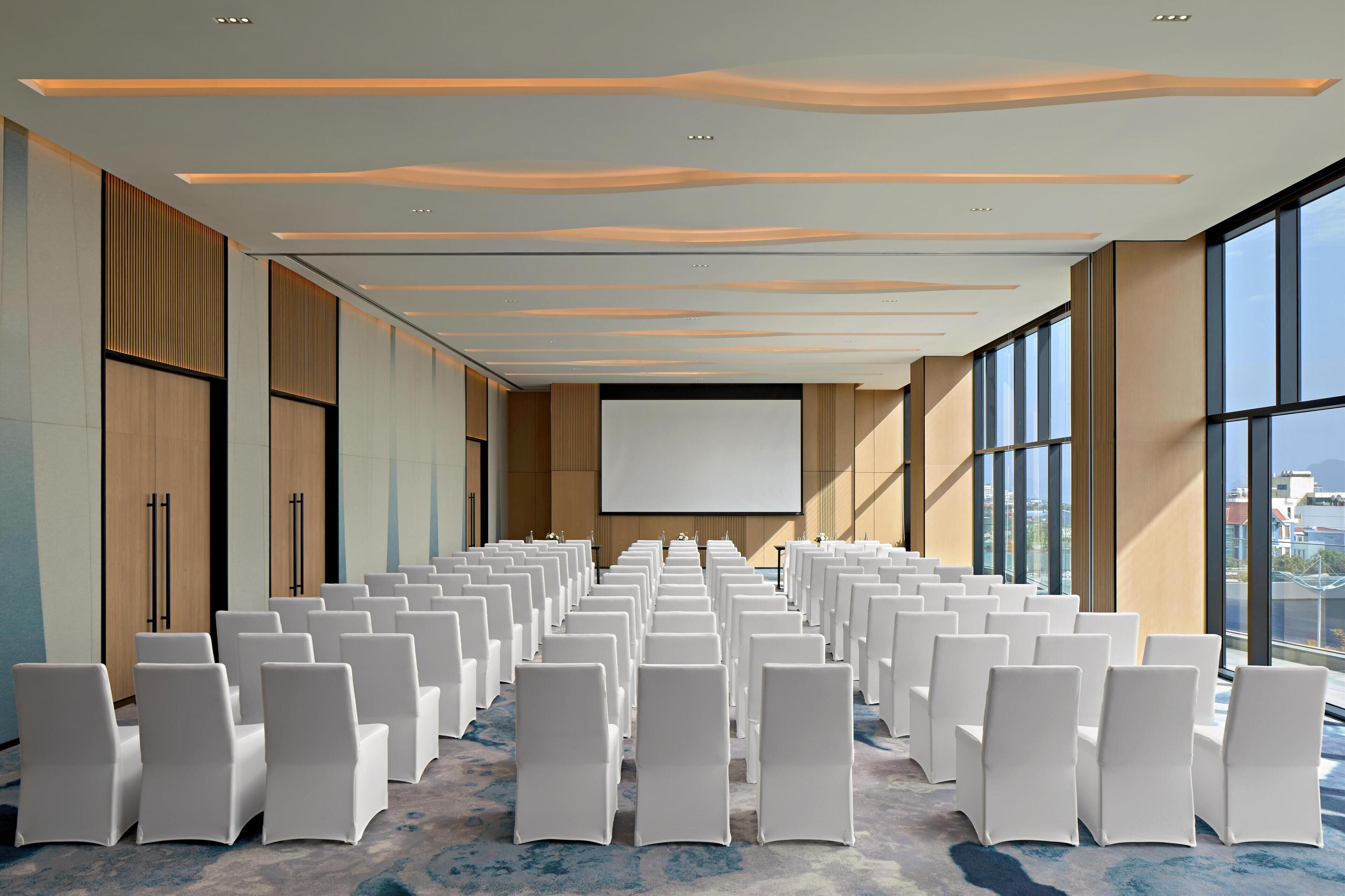 A conference room with rows of white chairs, a large screen, and a modern ceiling design.