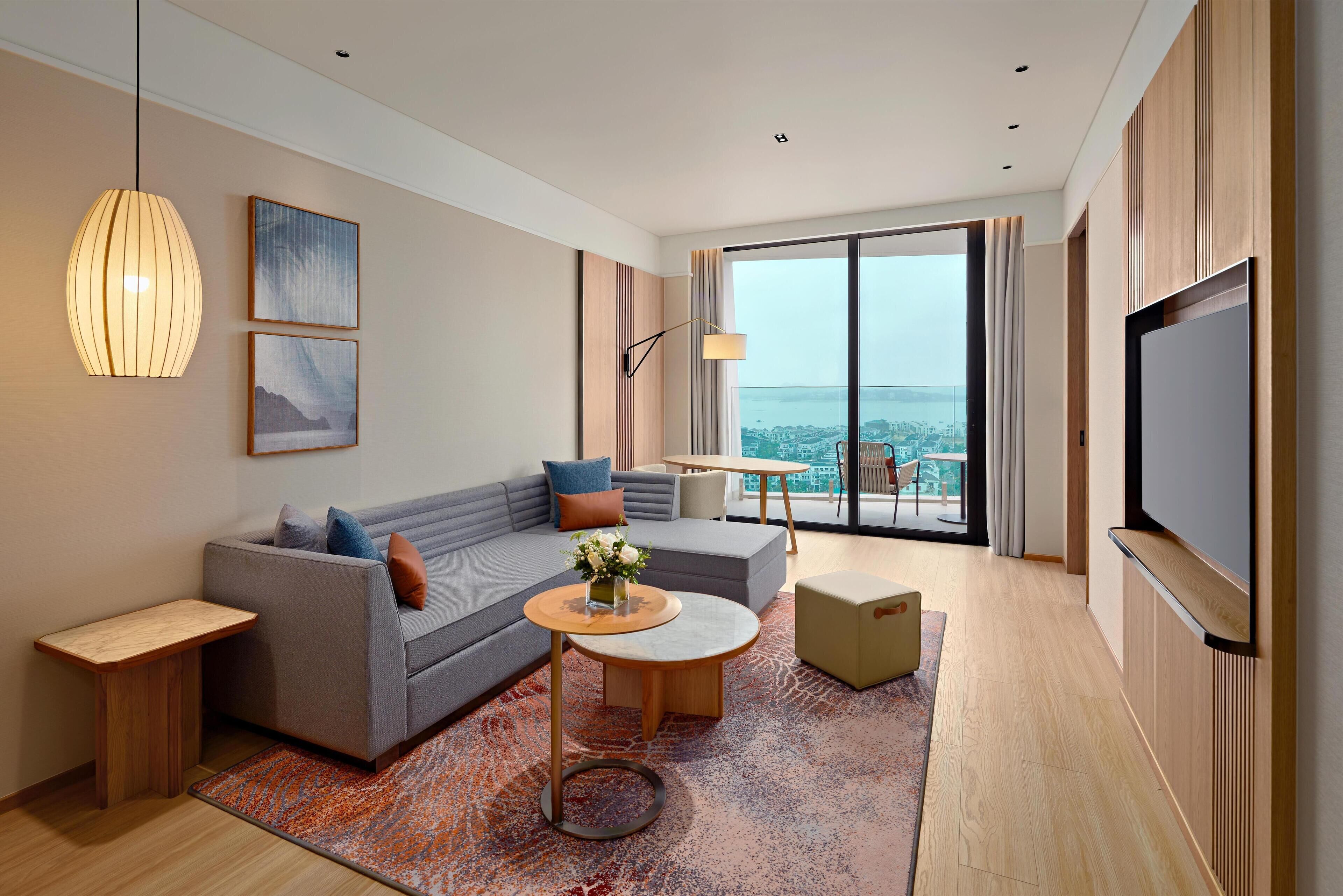 A modern living room with a sofa, coffee table, and TV. It features a large window with a city view.