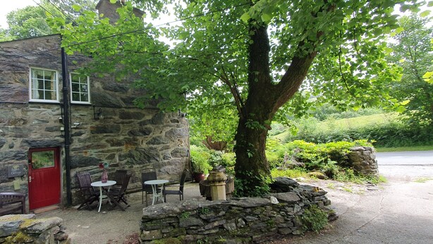A stone cottage with a red door, outdoor seating, and a large tree providing shade. - Sygun Cottage 40 mins walk to Watkin Waterfalls, Snowdon (Beddgelert)