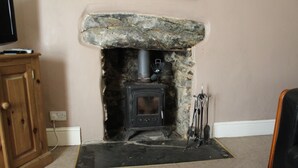 A stone fireplace with a black wood-burning stove, a wooden cabinet to the left, and a black leather chair to the right. - Sygun Cottage 40 mins walk to Watkin Waterfalls, Snowdon (Beddgelert)