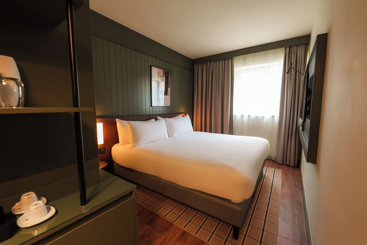A hotel room with a large bed, a bedside table, a lamp, and a TV mounted on the wall.