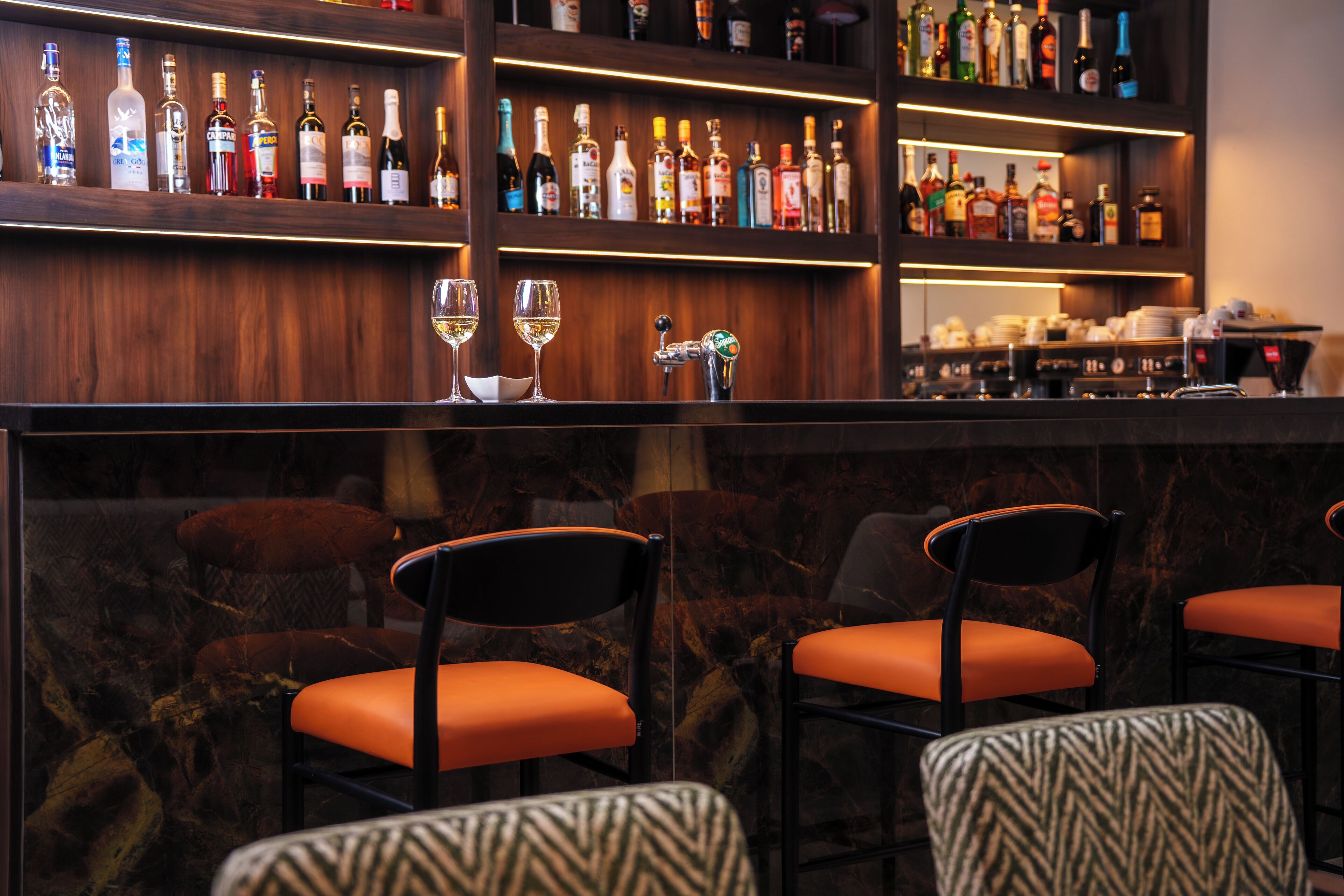 A bar area with shelves of liquor bottles, two glasses of white wine, and bar stools with orange cushions.