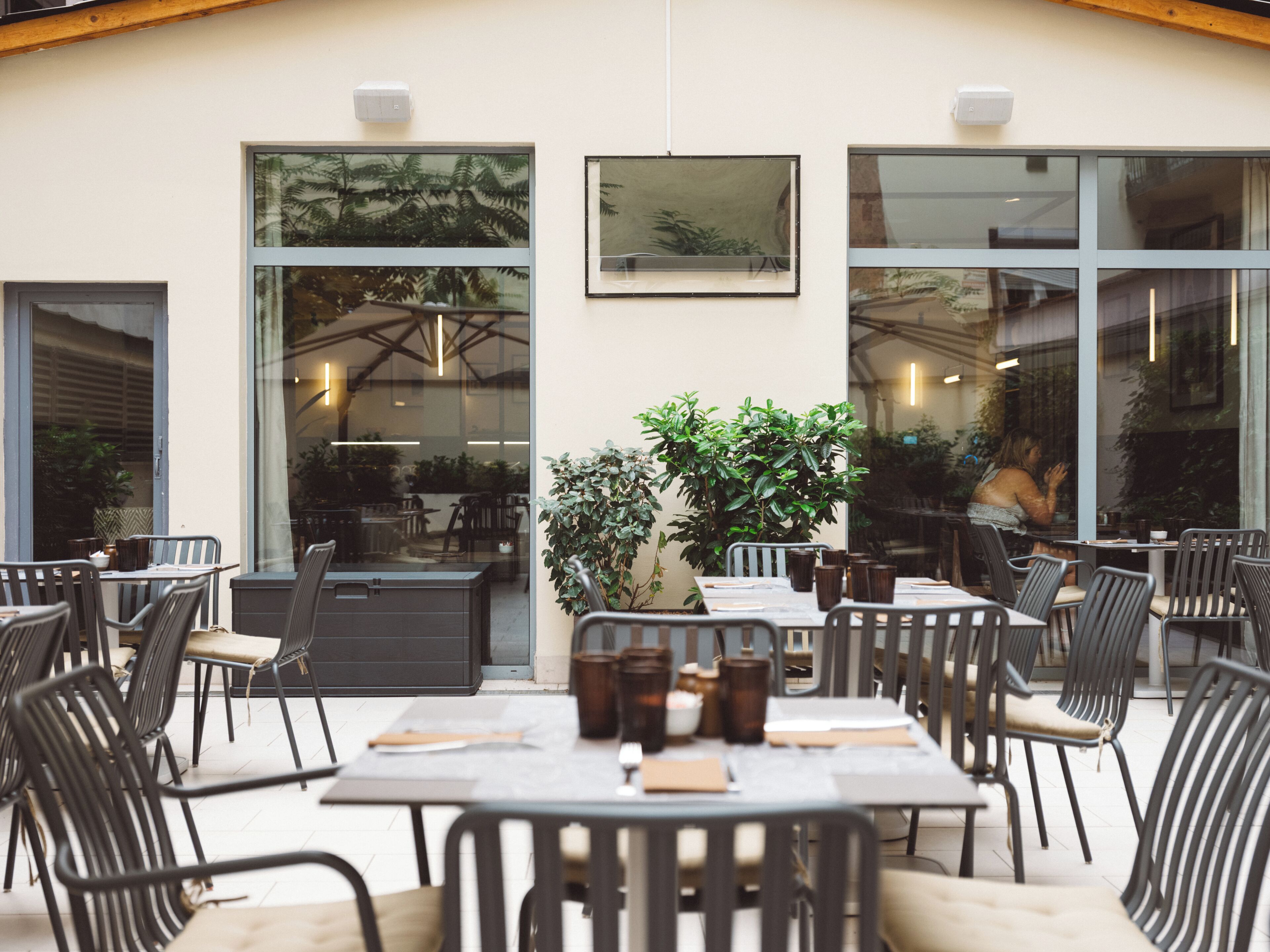 An outdoor dining area with tables and chairs, large windows, and potted plants.