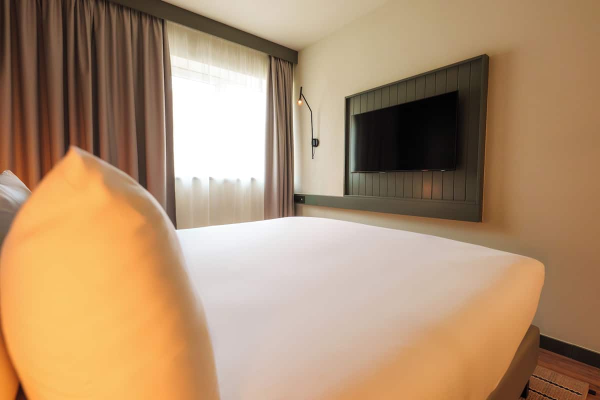 A hotel room with a large bed, a flat-screen TV mounted on the wall, and a window with curtains.
