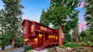 A two-story wooden cabin with a porch, surrounded by trees and a landscaped garden.