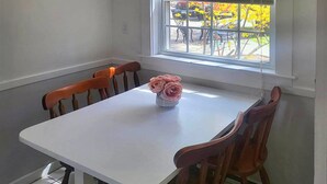 Een eethoek met een witte tafel en houten stoelen, een vaas met bloemen en een raam met zonwering.