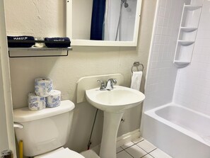 A bathroom with a toilet, sink, and bathtub. There are toilet paper rolls, a towel, and a shelf with makeup items.