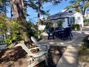 A patio with Adirondack chairs and a table, surrounded by trees and a white building with blue trim.